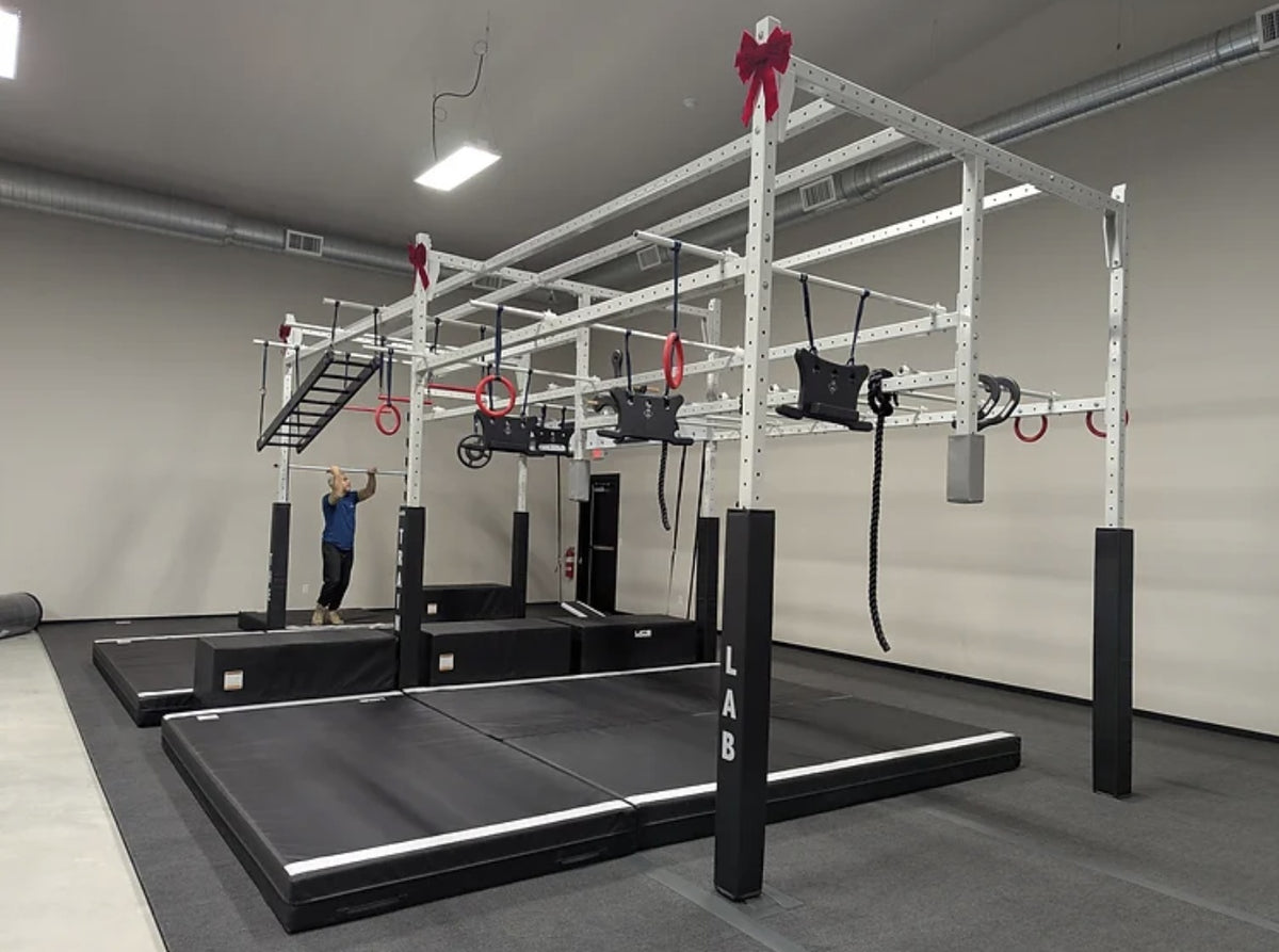 A large white and black functional strength training cage with gymnastic rings, pull-up bars, and hanging straps, set in a gym with black mats and a person visible in the background. Done by USA Finishing